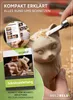 A close-up of a wooden cat carving being worked on by a hand with a chisel, with a booklet and a book cover in the foreground. Produktbild-Vorschau
