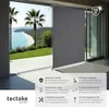 A grey vertical awning is shown partially deployed in front of a scenic view of a beach and palm trees. Náhled obrázku produktu