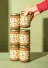 A hand is reaching for a stack of tahini jars against a green background. Produktbild-Vorschau