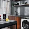 A bottle of AEG steam fragrance sits on a countertop next to a lemon and a small flower, with a washing machine in the background. Produktbild-Vorschau