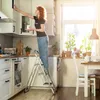 A woman is standing on a stepladder in a kitchen, reaching into a cabinet. Podgląd zdjęcia produktu
