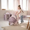 A young girl is playing with a doll stroller in a room with soft lighting and pink curtains. Náhled obrázku produktu