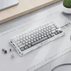 A white and gray mechanical keyboard is placed on a desk mat with a laptop and a potted plant in the background. Produktbild-Vorschau