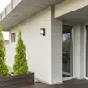 An outdoor wall light mounted on a white textured wall next to a window and some greenery. Podgląd zdjęcia produktu