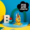 A colorful desk setup featuring a LEGO pencil holder shaped like a banana, a notebook, and other stationery items. Podgląd zdjęcia produktu