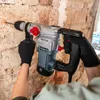 A person wearing black gloves is using a Practixx PX-BH-5 Bohrhammer to drill into a brick wall. Produktbild-Vorschau