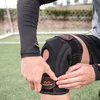 A person is adjusting a black knee brace with red accents on a green turf field. Podgląd zdjęcia produktu