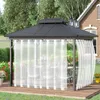 A gazebo with white sheer curtains set up on a wooden deck, with a table and chairs inside. The gazebo is surrounded by greenery and a brick house is visible in the background. Produktbild-Vorschau