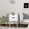 A white nightstand with two drawers sits next to a sofa. A plant is on the left, and books and a cup are on top of the nightstand. Produktbild-Vorschau