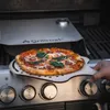 A pizza is being placed onto a grill using a pizza peel. Podgląd zdjęcia produktu
