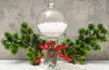 A clear glass Christmas ornament filled with artificial snow, placed on a stand decorated with pine sprigs, pinecones, red berries, and a red ribbon with "Merry Christmas" text. It is flanked by artificial pine branches against a textured gray background. Podgląd zdjęcia produktu