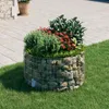 A gabion planter filled with plants sits on a lawn next to a paved area. Náhled obrázku produktu