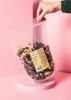 A hand holding a bag of student food (nuts and dried fruit) over a pink pedestal. Produktbild-Vorschau