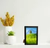 A black picture frame with a landscape image displayed inside, placed on a white shelf next to decorative vases and plants. Náhled obrázku produktu
