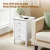 A white nightstand with three drawers is placed next to a cream-colored sofa, with a plant and a phone charging on top. Podgląd zdjęcia produktu