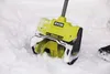 A close-up of a yellow and black electric snow blower with the Ryobi logo, partially covered in snow, sitting in a snowy environment. Produktbild-Vorschau