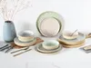 A collection of ceramic dinnerware, including plates and bowls, arranged on a white surface with a light gray background. A vase with dried branches is on the left. Produktbild-Vorschau