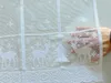 A hand holding a white lace curtain with a Christmas-themed pattern of deer and snowflakes. Produktbild-Vorschau