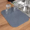 A gray silicone drying mat with a wavy pattern is placed on a wooden countertop next to a stainless steel sink. Two clear glass cups with handles are placed upside down on the mat. Produktbild-Vorschau