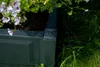 A close-up view of a dark gray planter box with flowers growing in it, set against a background of green grass and foliage. Produktbild-Vorschau