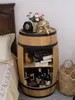 A barrel-shaped mini bar stand next to a bed, decorated with a mask and flowers on top, and bottles and books inside. Produktbild-Vorschau
