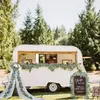 A white vintage caravan converted into a bar, decorated with greenery and fabric, set up outdoors in a grassy area with trees in the background. Anteprima dell’immagine prodotto