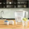 A water filter pitcher and replacement cartridges are shown on a wooden countertop in a kitchen setting. Produktbild-Vorschau