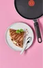 A crepe with chocolate sauce and bananas on a plate, next to a spoon and a frying pan, set against a pink background. Produktbild-Vorschau