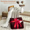 A dark red sherpa blanket is folded and tied with a red ribbon, presented as a gift. A lighter colored sherpa blanket is draped over a wooden chair in the background. A woven basket with a blanket inside and a stack of books are also visible. Podgląd zdjęcia produktu