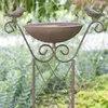 A close-up view of a rustic bird bath with two decorative birds on a stand, set against a blurred green garden background. Náhled obrázku produktu