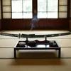A katana sword is displayed on a stand on a low table, with small cups and an incense burner. Smoke rises from the incense. Produktbild-Vorschau