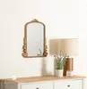 A decorative gold mirror is mounted on a white wall above a light wood dresser. A table lamp and a potted plant are on the dresser. Anteprima dell’immagine prodotto