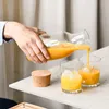A hand is pouring orange juice from a glass carafe into two glasses filled with ice. Produktbild-Vorschau