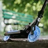 A close-up of a black scooter with blue LED wheels, resting on a path with a blurred green bench and trees in the background. Voir les images du produit