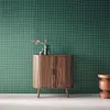 A wooden sideboard with decorative doors stands against a green grid-patterned wall. A small orange stool is to the right. There are two vases on top of the sideboard. Produktbild-Vorschau