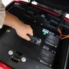 A hand is shown reaching into the storage compartment of a red three-wheeled vehicle, likely to access or remove the battery. Produktbild-Vorschau