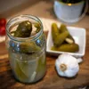 A close-up shot of a glass jar filled with pickles, alongside a small bowl of pickles and a head of garlic on a wooden surface. Produktbild-Vorschau