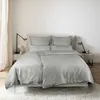A neatly made bed with gray satin bedding, set against a white wall with a sheer curtain on the left and two bedside tables with lamps. Produktbild-Vorschau