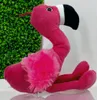 A plush flamingo toy is shown sitting in front of a green leafy background and a white wall. Produktbild-Vorschau