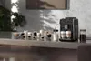 A coffee machine is shown on a counter with several glasses of coffee and milk, and a jar of coffee beans. Produktbild-Vorschau