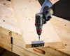 A person is using a cordless drill to screw a hinge onto a wooden board, which is clamped to a workbench. Produktbild-Vorschau