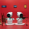 Two robots are depicted in a gallery-like setting with paintings on a red wall. One robot is painting on an easel, and the other is playing a violin. Náhled obrázku produktu