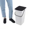 A person's legs and feet are shown next to a white trash can with a black lid and handle. Produktbild-Vorschau