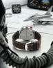 A watch with a brown leather strap is placed on a table with aviation-themed items like a model airplane and pilot helmet. Náhled obrázku produktu