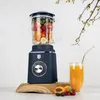 A navy blue stand mixer filled with fruit sits on a wooden board, next to a glass of orange juice. Produktbild-Vorschau