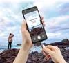 A person is holding a smartphone that displays a "Cannot Take Photo" message, while simultaneously plugging a USB stick into the phone. Another person stands in the background on a rocky shore overlooking the ocean. Anteprima dell’immagine prodotto