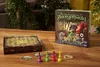 A board game box and its components are displayed on a wooden table. Produktbild-Vorschau
