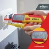 A hand is holding a voltage tester and inserting it into a wall socket. A close-up inset shows the tester's display. Náhľad obrázku produktu