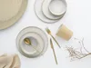 A flat lay of a 16-piece stoneware dinnerware set in white sand color, with gold cutlery and a beige paper cup, arranged on a white background. Produktbild-Vorschau