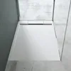 A modern white shower tray with a linear drain, set against a concrete floor and a grey wall, with glass shower enclosure panels visible. Produktbild-Vorschau